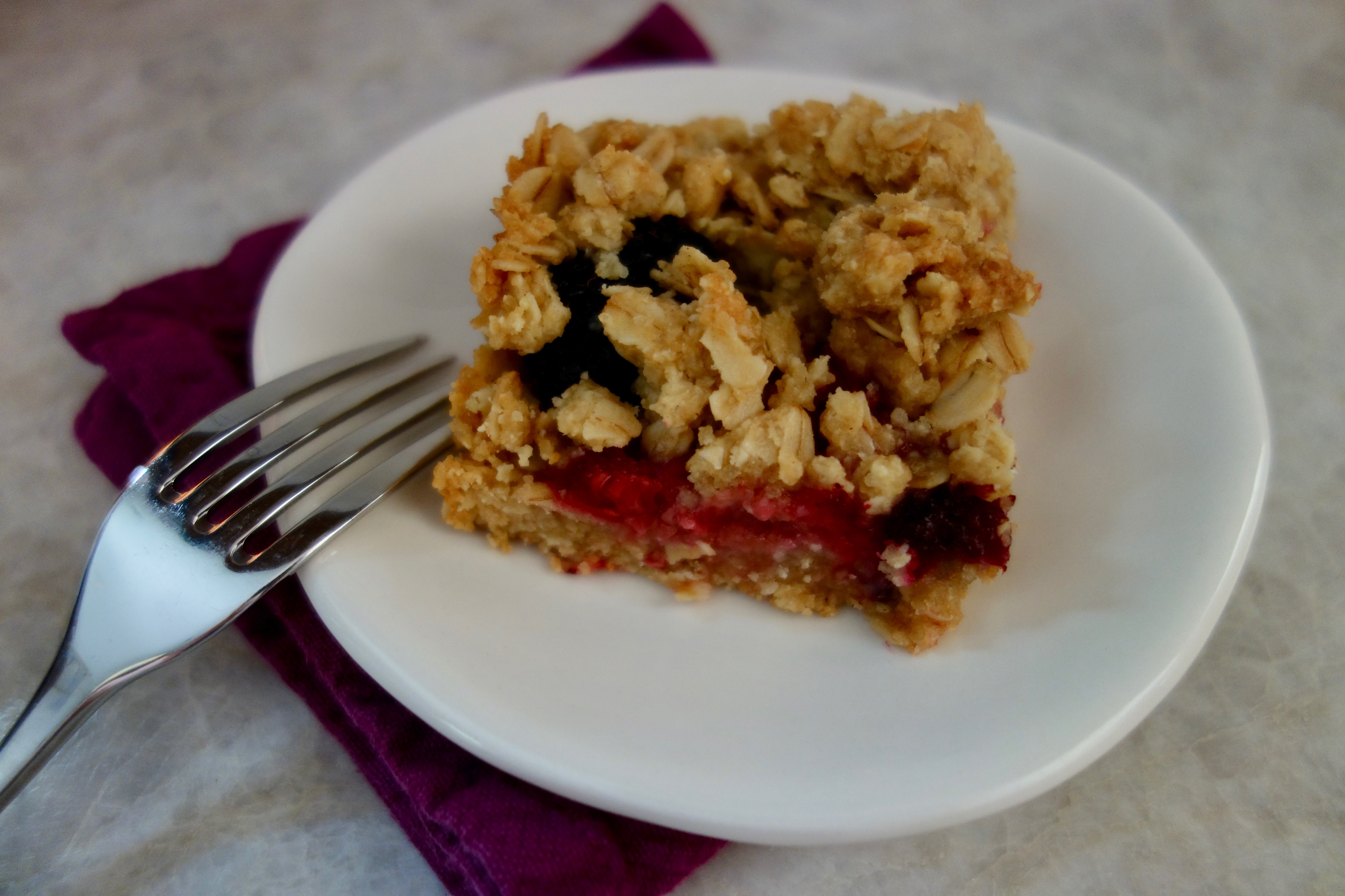 single square of oatmeal crumble berry bar on plate with fork and purple napkin