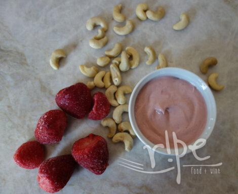 Ramekin of pink strawberry cashew cream surrounded by 6 frozen strawberries and a scatter of raw cashews on stone countertop